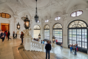 Ornate palace interior with grand staircase and many visitors.