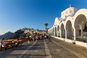 Crowds walk past white church cliffside town in Santorini.
