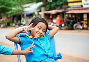 Smiling girl in blue makes peace sign outdoors.
