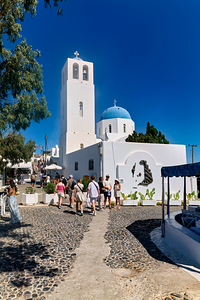 Greek village street with white church blue dome and tourists.