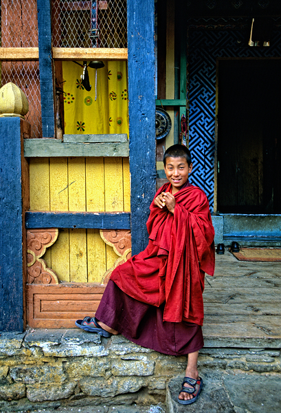 Smiling young monk in red robes at colorful traditional building by Marco Brivio