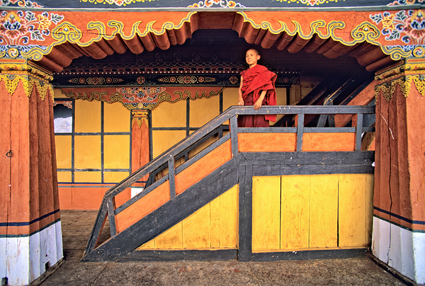 Young monk in red robes on colorful temple staircase. by Marco Brivio