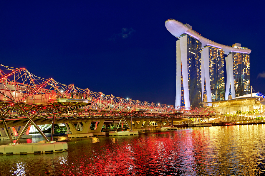 People walk across the Helix Bridge as the Marina Bay Sands Hotel shines at sunset. The sky turns orange and lights reflect in the water.  Print