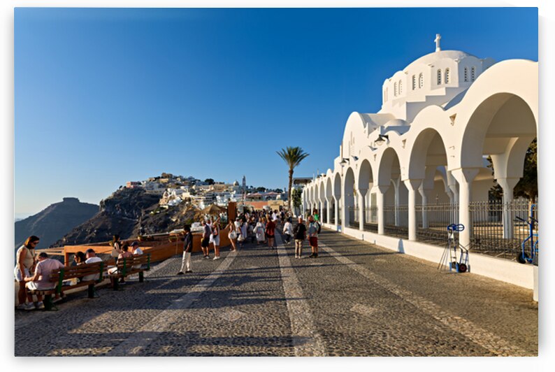 Crowds walk past white church cliffside town in Santorini. by Marco Brivio