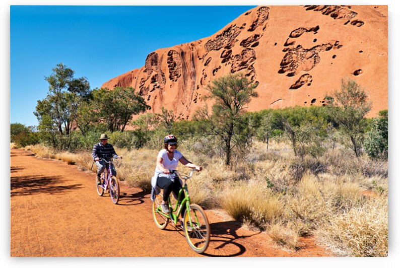 Two people cycling near Uluru under a clear blue sky. by Marco Brivio