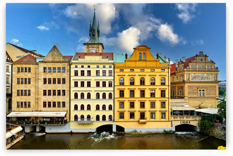 Historic Prague buildings and clock tower line a scenic river. by Marco Brivio