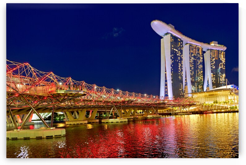 People walk across the Helix Bridge as the Marina Bay Sands Hotel shines at sunset. The sky turns orange and lights reflect in the water. by Marco Brivio