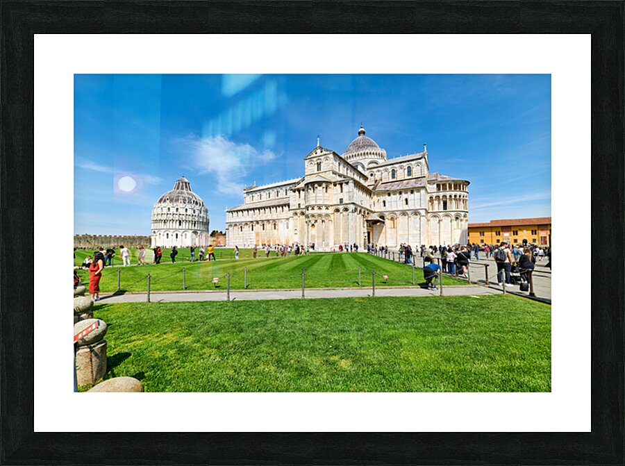People walk in Piazza dei Miracoli admiring the Baptistry and Cathedral in Pisa. The sun shines on the historic buildings and green grass. Picture Frame print