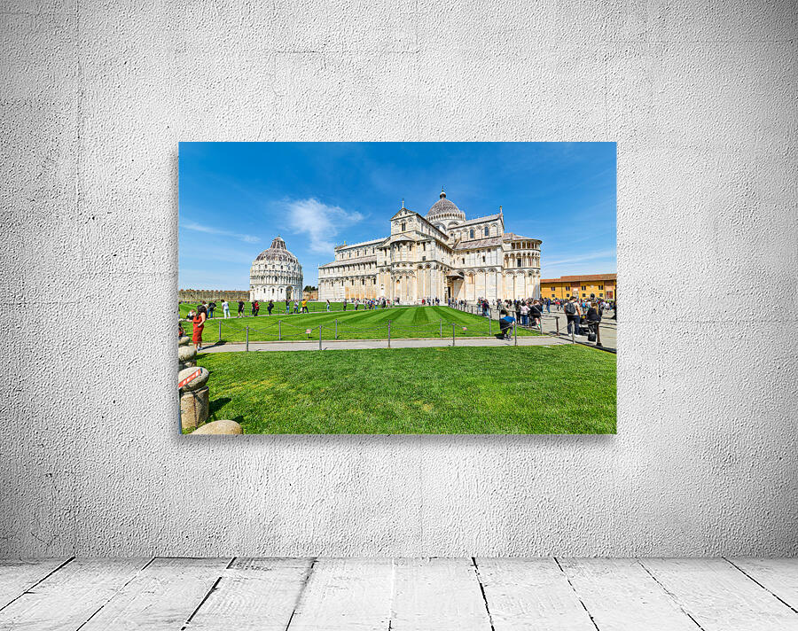 People walk in Piazza dei Miracoli admiring the Baptistry and Cathedral in Pisa. The sun shines on the historic buildings and green grass. Wall Preview