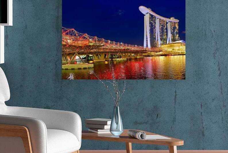 People walk across the Helix Bridge as the Marina Bay Sands Hotel shines at sunset. The sky turns orange and lights reflect in the water. by Marco Brivio