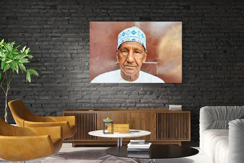 Elderly man in traditional dress sits by brown wall by Marco Brivio