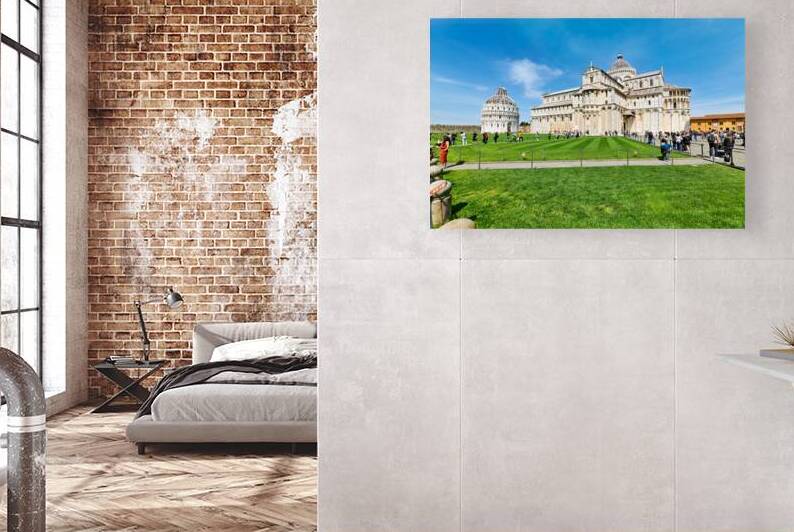 People walk in Piazza dei Miracoli admiring the Baptistry and Cathedral in Pisa. The sun shines on the historic buildings and green grass. by Marco Brivio
